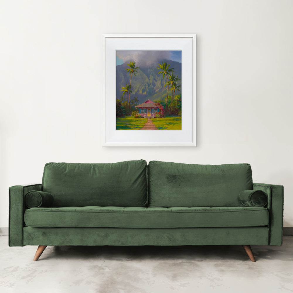 A Hawaiian painting is shown as a framed wall art print hanging on a living room wall over a green couch. The artwork shows a scene from Hanalei on the island of Kauai and was painted by artist Karen Whitworth.