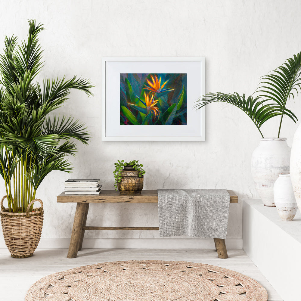 Bird of paradise wall art print of 2 tropical flowers with leaves and foliage in the background. The artwork is framed in a white picture frame in a tropical home interior.