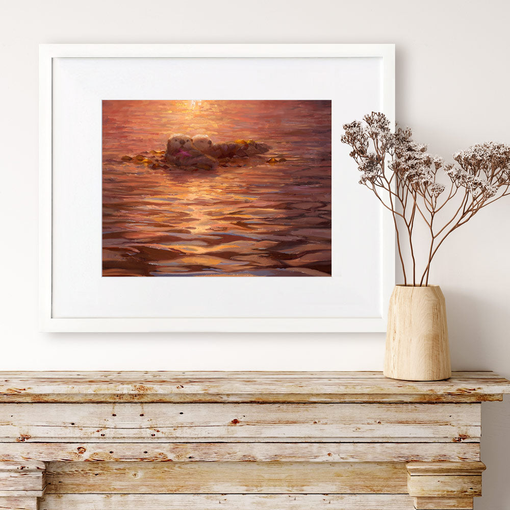 Sea otter wall art print of an ocean sunset painting featuring cute animals. The artwork is framed in a white wood picture frame and is hanging above a rustic farmhouse fireplace mantel.