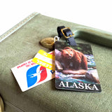 An Alaska luggage tag rests on a vintage suitcase with other tourist and travel tags. The luggage tag depicts a brown bear and features a leather buckle strap.