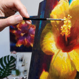 An artist's hand holding a paintbrush applies paint to a large tropical wall art canvas of yellow hibiscus flowers