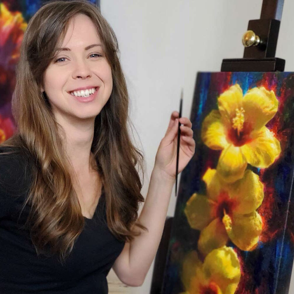 Artist Karen Whitworth in her Studio with a brightly colored tropical hibiscus painting on canvas resting on an easel. The artist is holding a paintbrush and smiling at the camera.