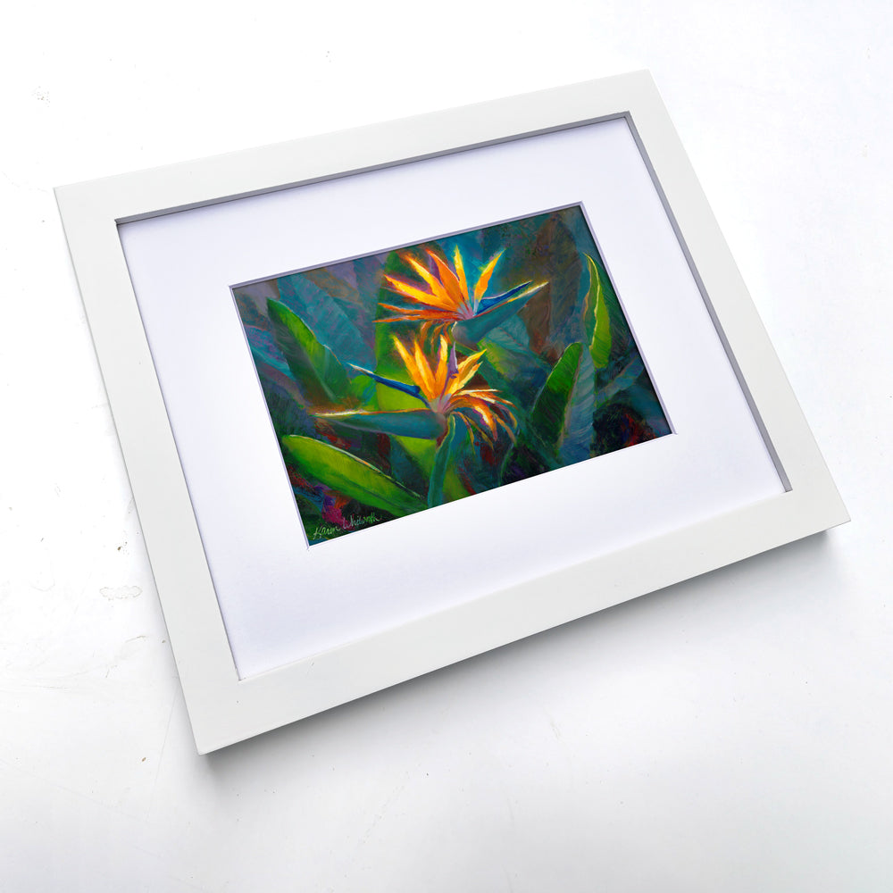 Bird of paradise wall art print of 2 tropical flowers with leaves and foliage in the background. The artwork is framed in a white picture frame on a white table.