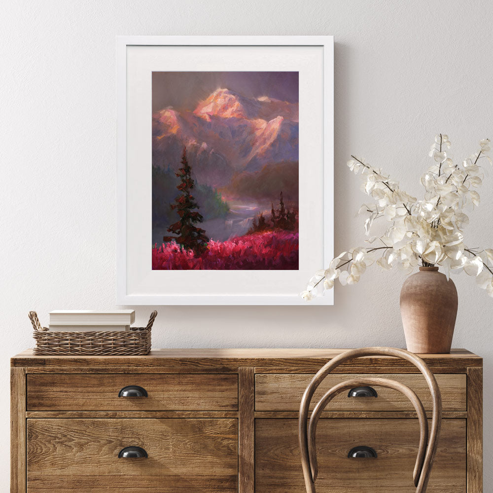 Alaska wall art print of a mountain sunset landscape painting depicting Denali and a field of fireweed wildflowers. The painting is titled "Denali Summer" by Karen Whitworth and is framed in a white wood picture frame. It is hanging on a white wall behind a rustic farmhouse home decor dresser.