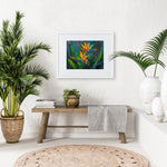 Bird of paradise wall art print of 2 tropical flowers with leaves and foliage in the background. The artwork is framed in a white picture frame in a tropical home interior.