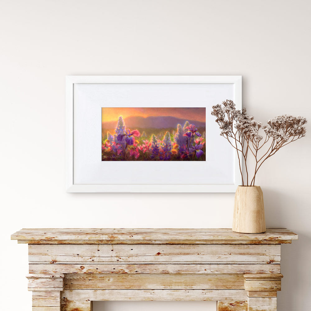 Framed wildflower wall art print featuring a flower field landscape painting of Sleeping Lady Mountain, also known as Mt Susitna, in Southcentral Alaska. The artwork is framed in a white picture frame made of wood, and is hanging on a beige wall over a rustic farmhouse decor fireplace.