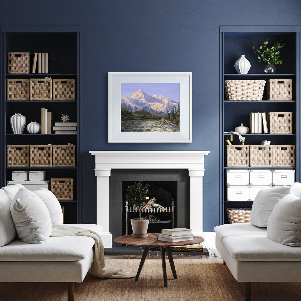 Alaska art print of Denali mountain wall art in a nature landscape painting. The artwork is displayed in a white wood picture frame over a fireplace mantel on a dark blue wall. The foreground of the scene shows a traditional living room with timeless home decor including a blue and white color scheme.