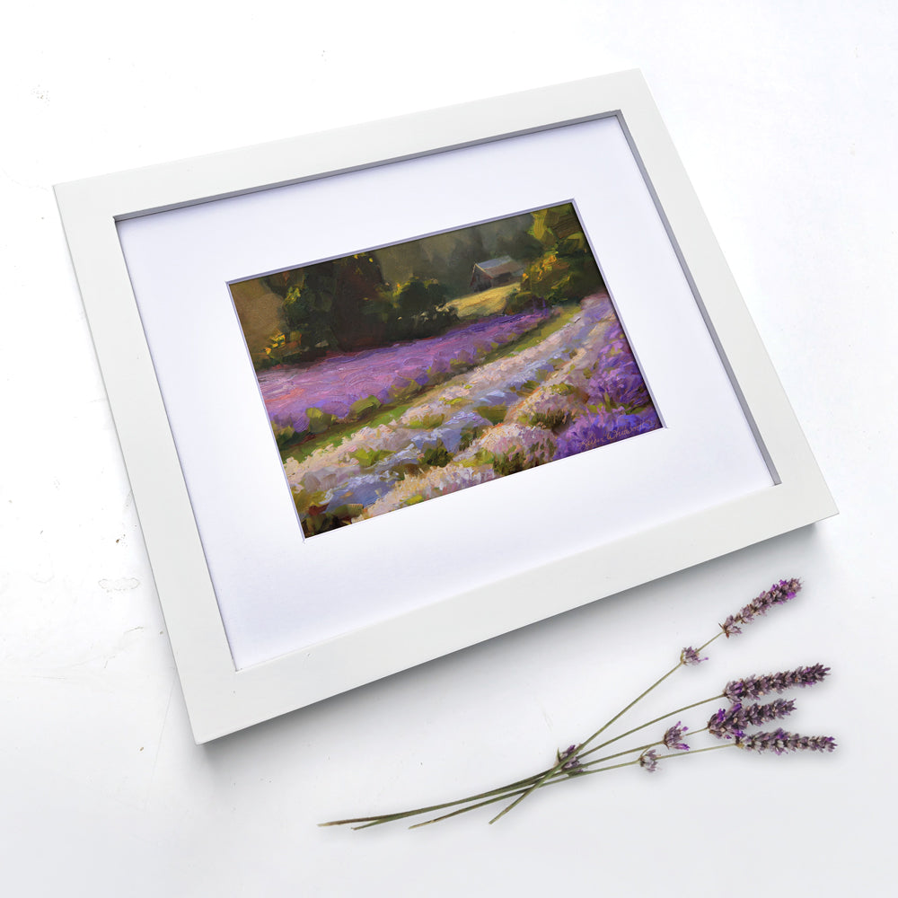Framed lavender field wall art painting featuring a rural farmhouse landscape at sunset. In the background a rustic barn stands against a forest of green trees. The artwork is framed in a white wood picture frame and is laying on a white table with blooming lavender stalks resting nearby.