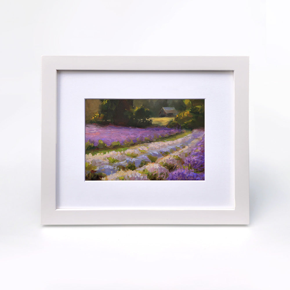 Framed lavender field wall art painting featuring a rural farmhouse landscape at sunset. In the background a rustic barn stands against a forest of green trees. The artwork is framed in a white wood picture frame and is hanging on a white wall.