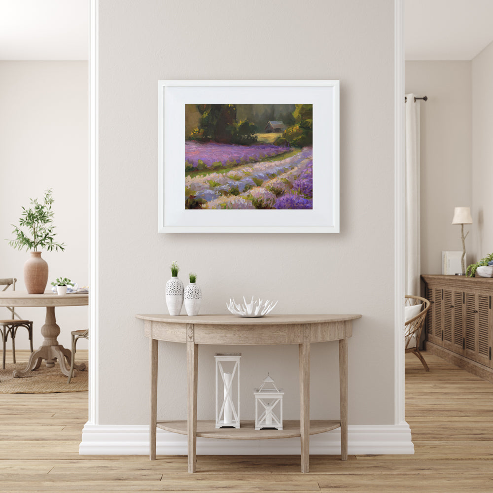 Framed lavender field wall art painting featuring a rural farmhouse landscape at sunset. In the background a rustic barn stands against a forest of green trees. The artwork is framed in a white wood picture frame and is hanging on a beige wall in a room with farmhouse decor and rustic natural wood dining table, console table, and hutch.