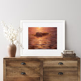 Sea otter wall art print of an ocean sunset painting featuring cute animals. The artwork is framed in a white wood picture frame and is hanging above a rustic farmhouse dresser.. 