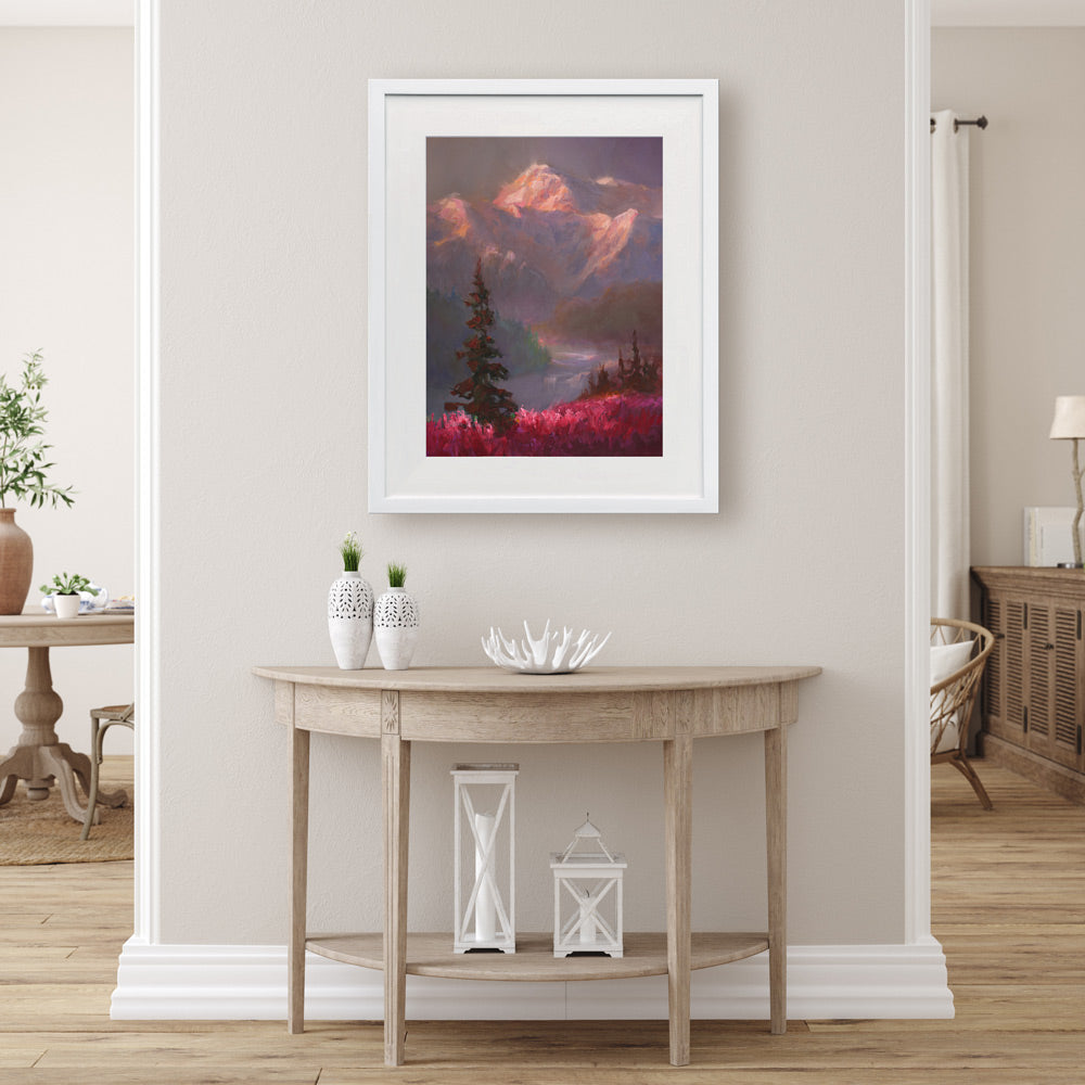 Alaska wall art print of a mountain sunset landscape painting depicting Denali and a field of fireweed wildflowers. The painting is titled "Denali Summer" by Karen Whitworth and is framed in a white wood picture frame. It is hanging on a beige wall behind a rustic farmhouse home decor table.