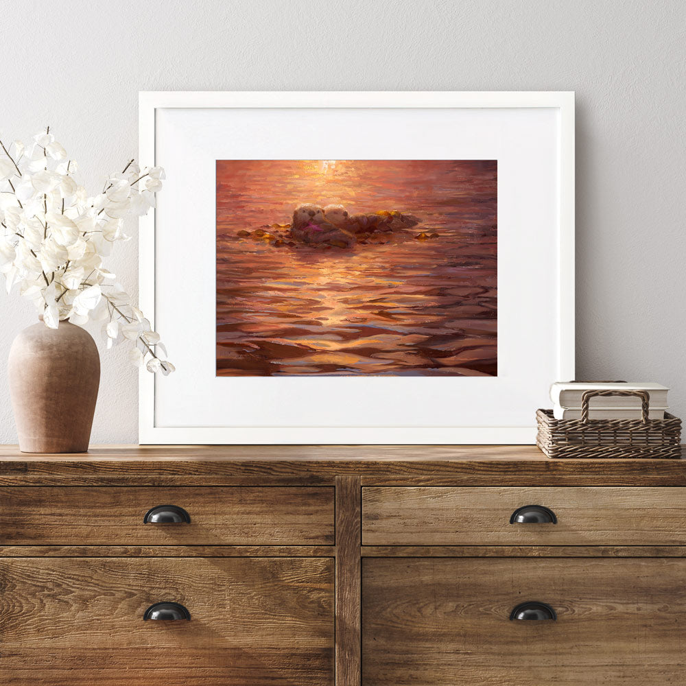 Sea otter wall art print of an ocean sunset painting featuring cute animals. The artwork is framed in a white wood picture frame and is hanging above a rustic farmhouse dresser.