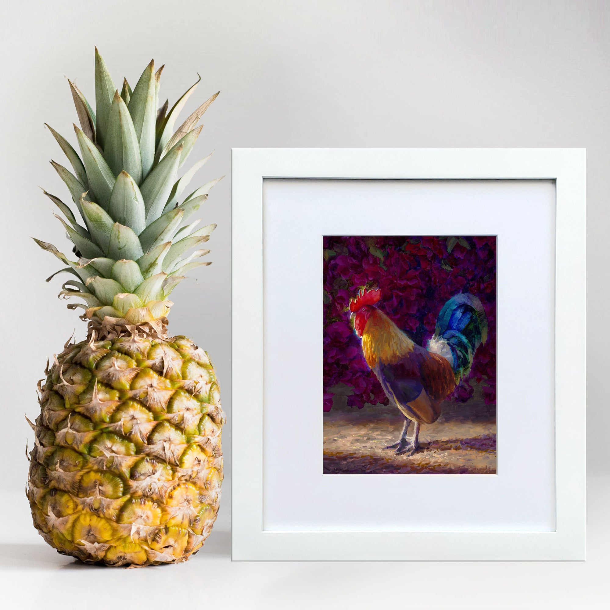 Rooster decor art print of a painting depicting a male chicken in front of a flowering pink bush. The wall art is framed in a white wood frame and standing in next to a pineapple.