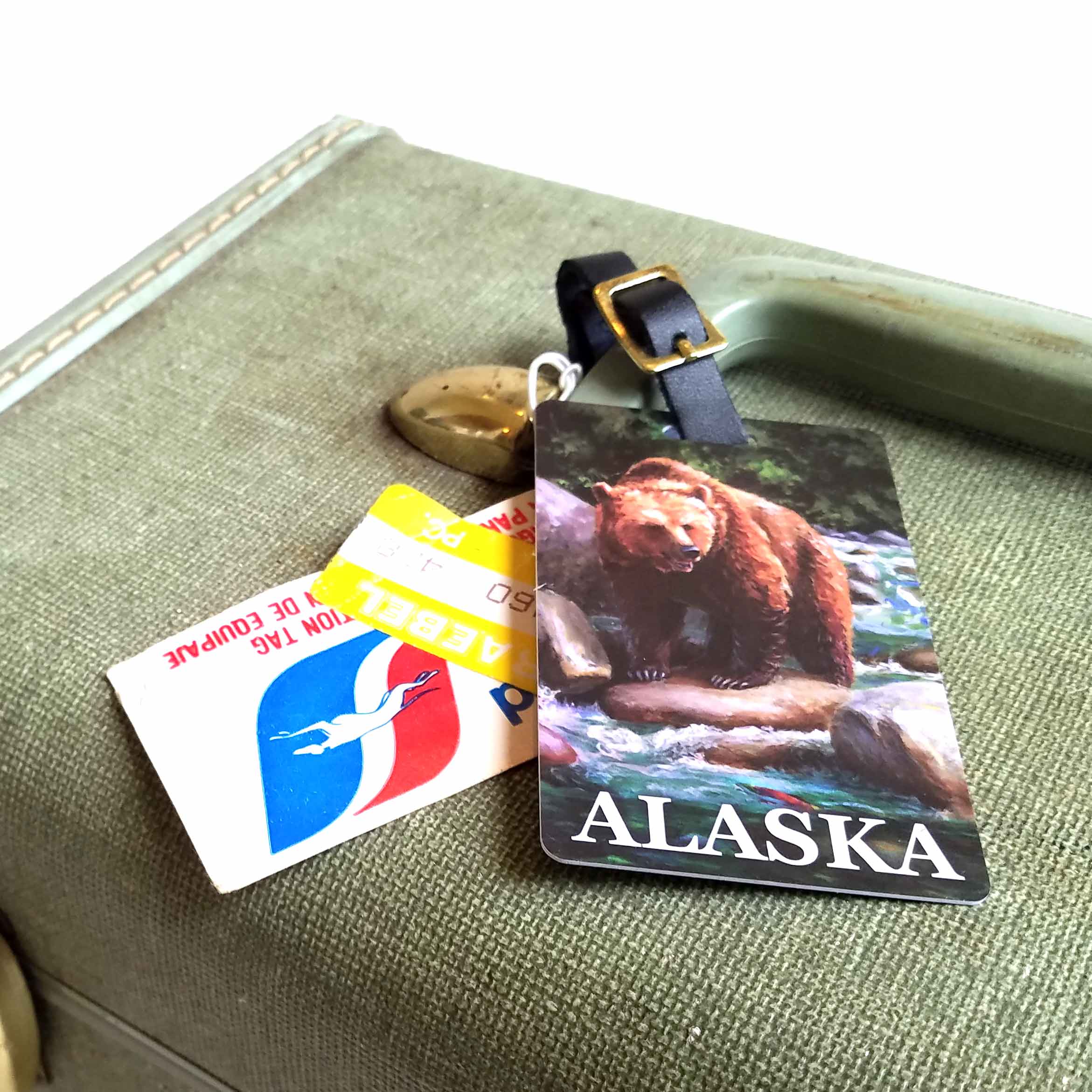 An Alaska luggage tag rests on a vintage suitcase with other tourist and travel tags. The luggage tag depicts a brown bear and features a leather buckle strap.