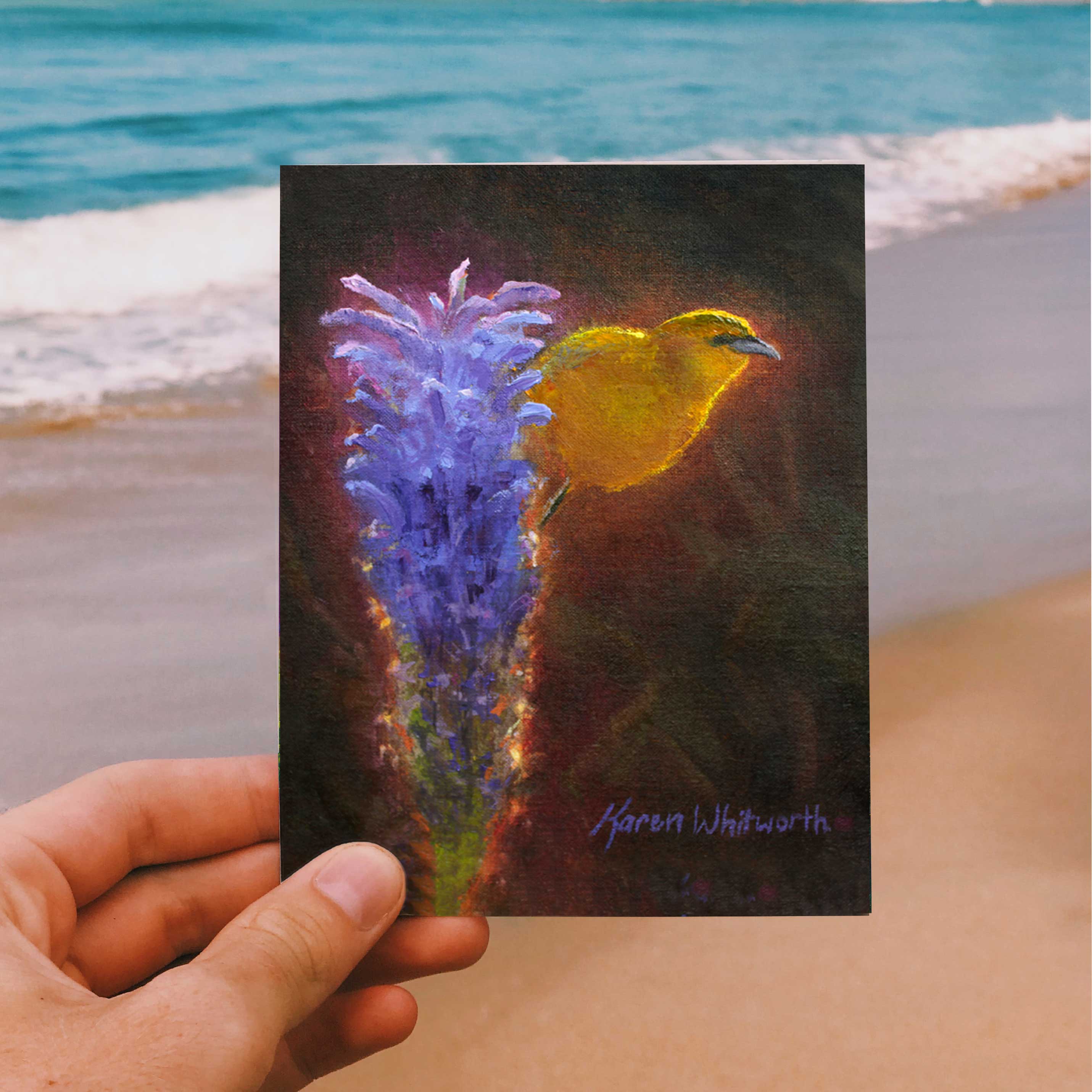 Person holding a Hawaii note card featuring painting of Amakihi Bird and Haleakala Lobelia Flower against a beach background