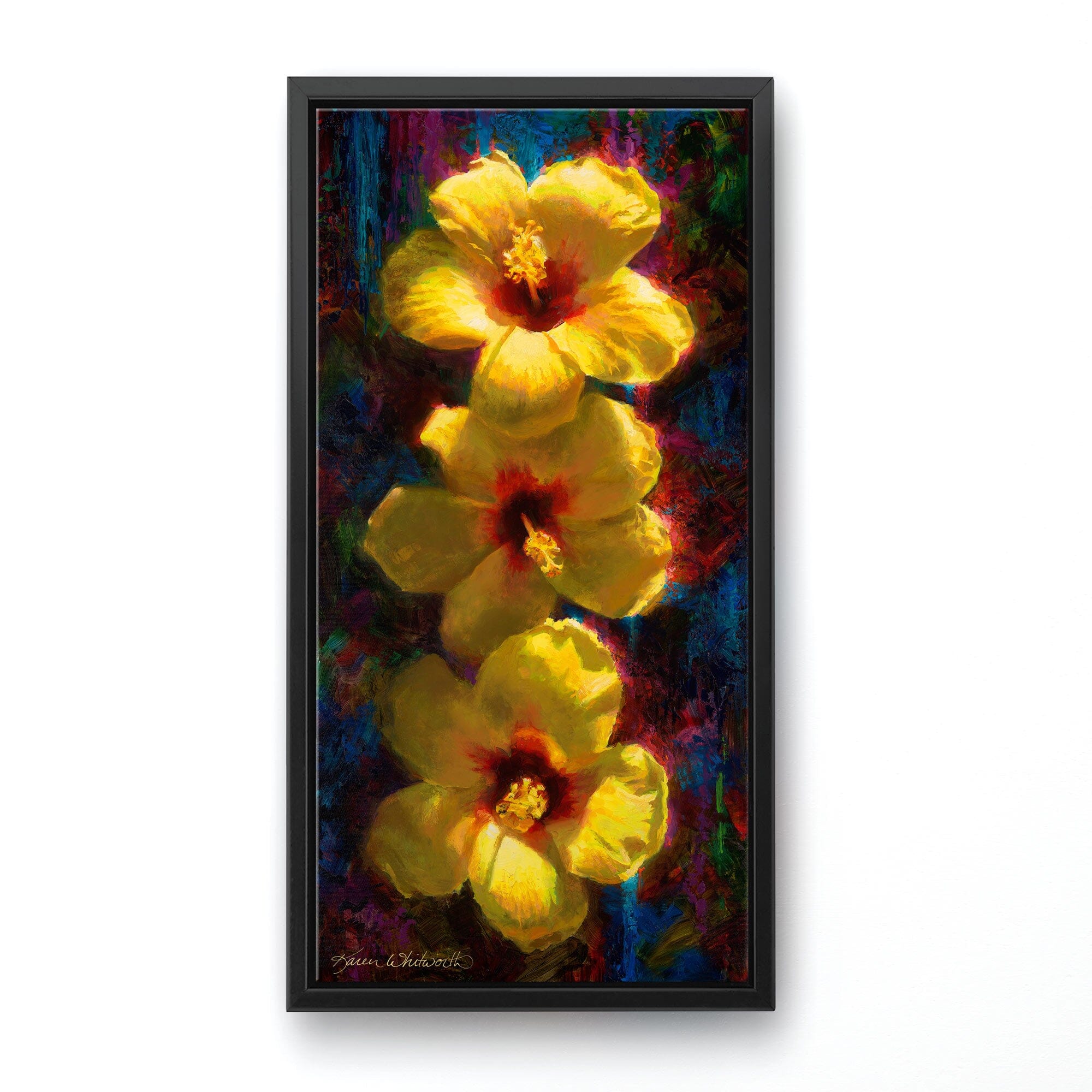 Vertical floral painting of tropical hibiscus flowers by artist Karen Whitworth. The 3 bright yellow flowers sit one on top of the other illuminated in sunlight against a dark background. The painting is framed in a sleek black contemporary frame and hanging on a white wall. 
