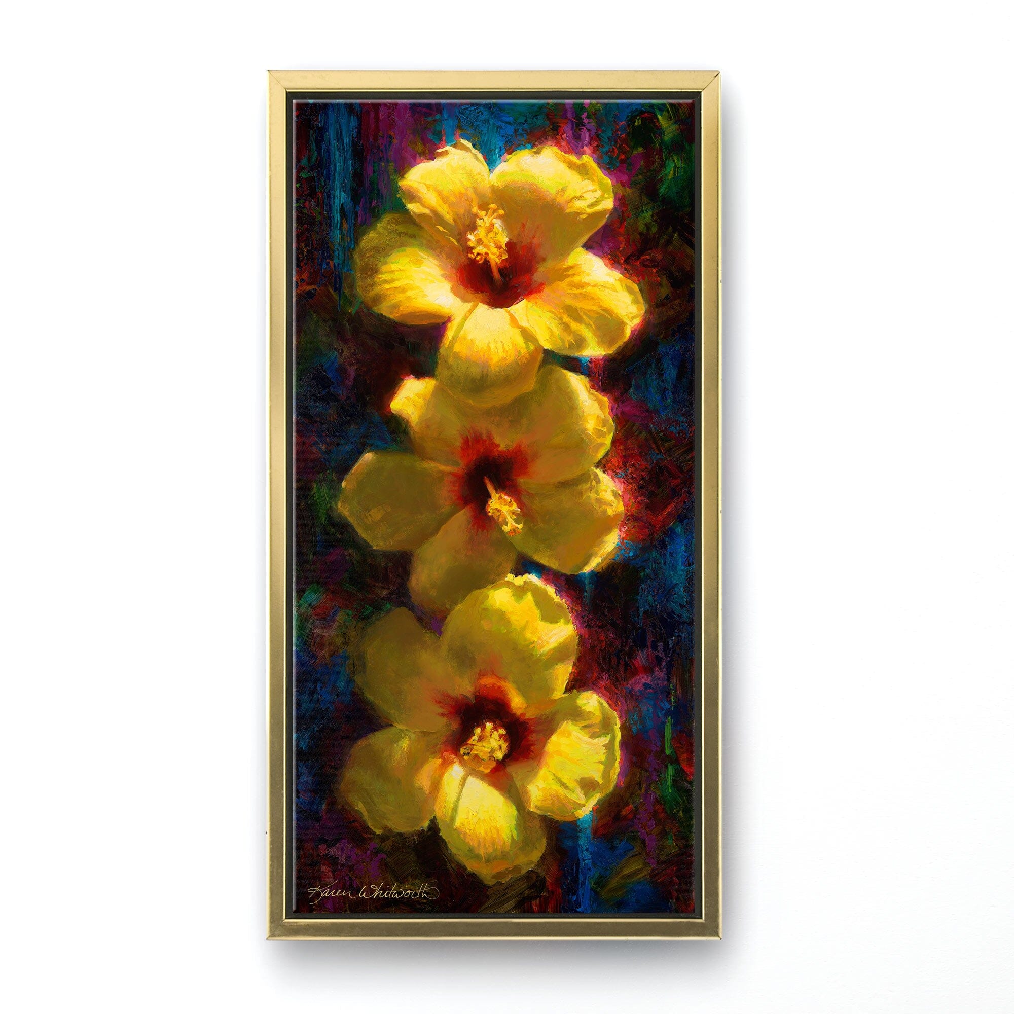 Vertical floral painting of tropical hibiscus flowers by artist Karen Whitworth. The 3 bright yellow flowers sit one on top of the other illuminated in sunlight against a dark background. The painting is framed in a sleek gold contemporary frame and hanging on a white wall. 
